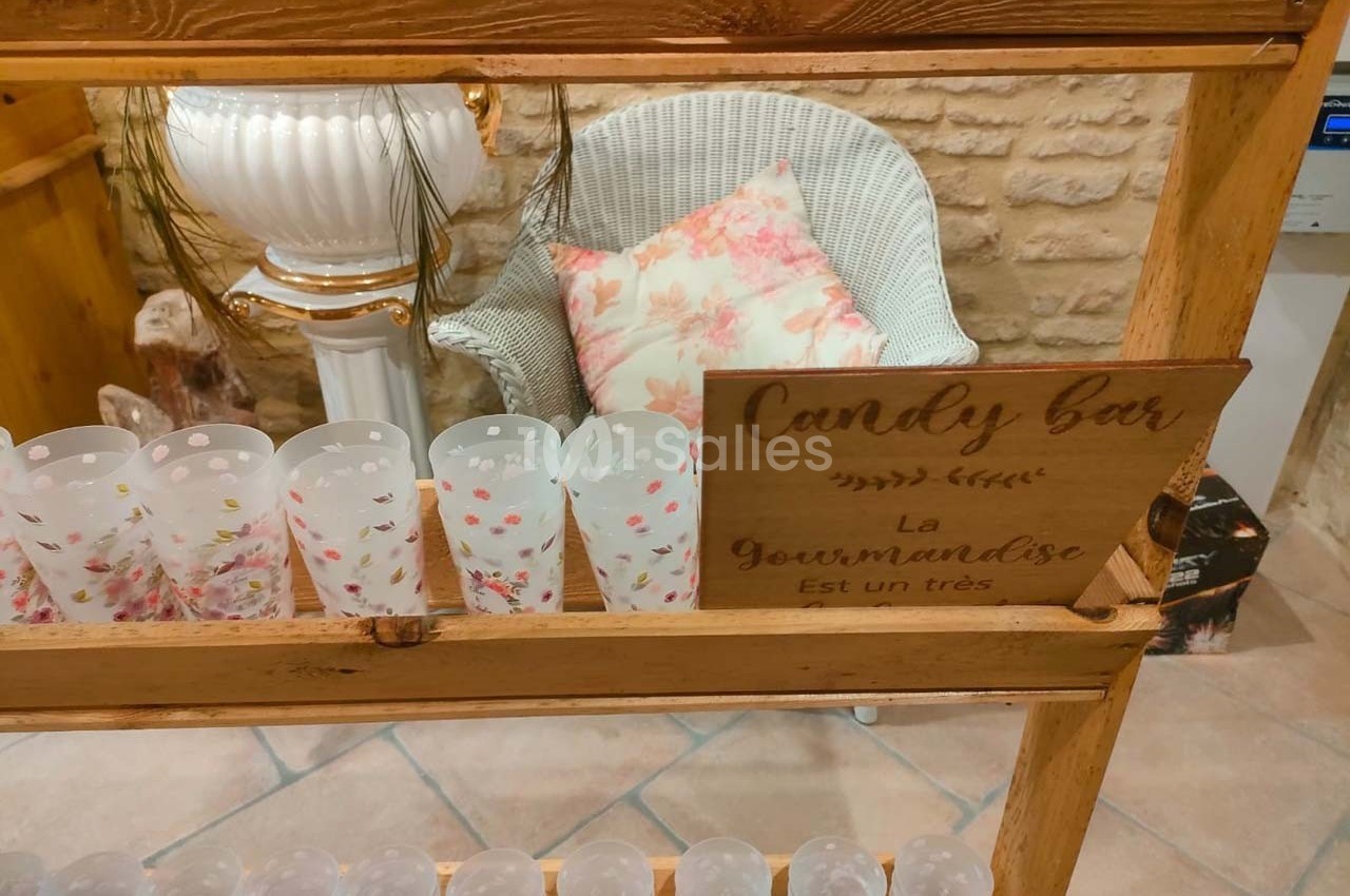 Étagère en bois avec des gobelets décorés, un fauteuil en osier et une pancarte ’Candy bar’ dans un décor rustique.