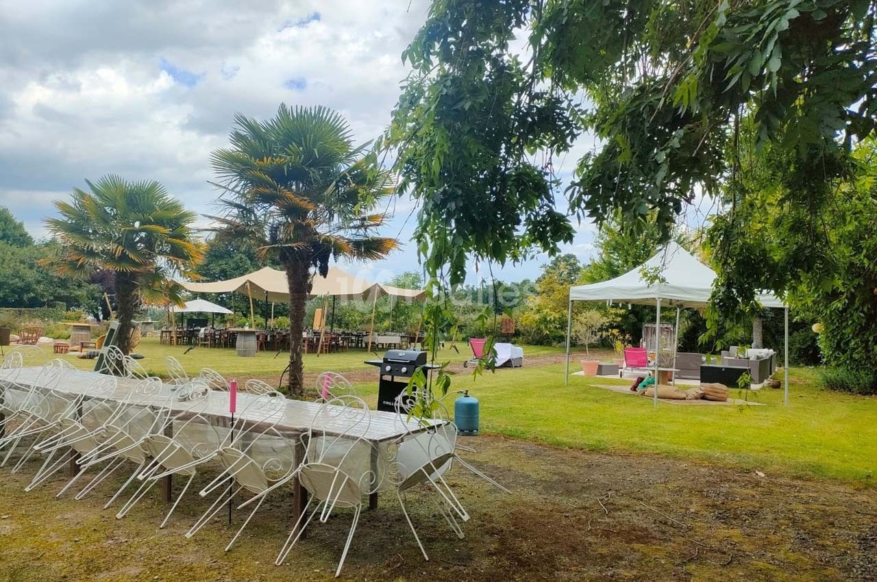 Tables et chaises blanches disposées en extérieur sous des arbres, avec des tentes et un espace aménagé dans un jardin.
