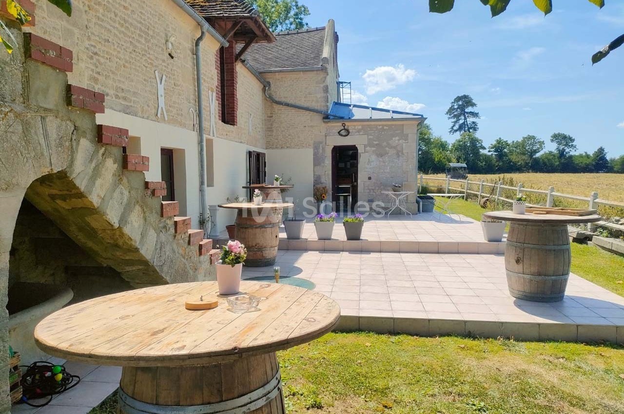 Terrasse ensoleillée avec tables en tonneaux, fleurs en pot et vue sur un champ bordé d'arbres.
