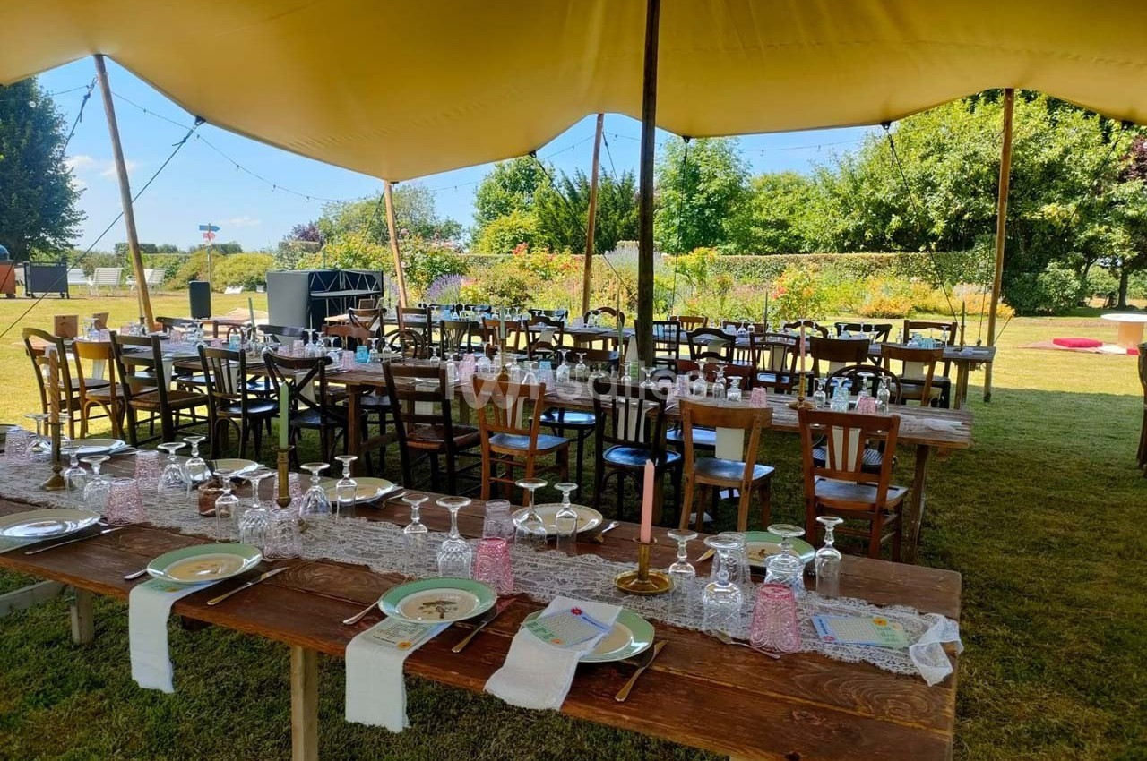 Tables en bois décorées pour un repas en plein air sous une tente, avec vaisselle et nappes en dentelle.