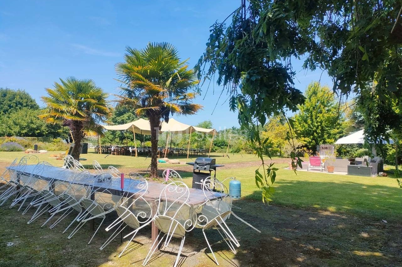 Tables et chaises blanches disposées en extérieur sous des arbres et des parasols, dans un jardin ensoleillé.