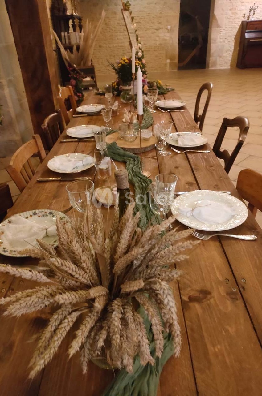 Table en bois décorée pour un repas, avec vaisselle, verres, bougies et un centre de table en épis de blé.