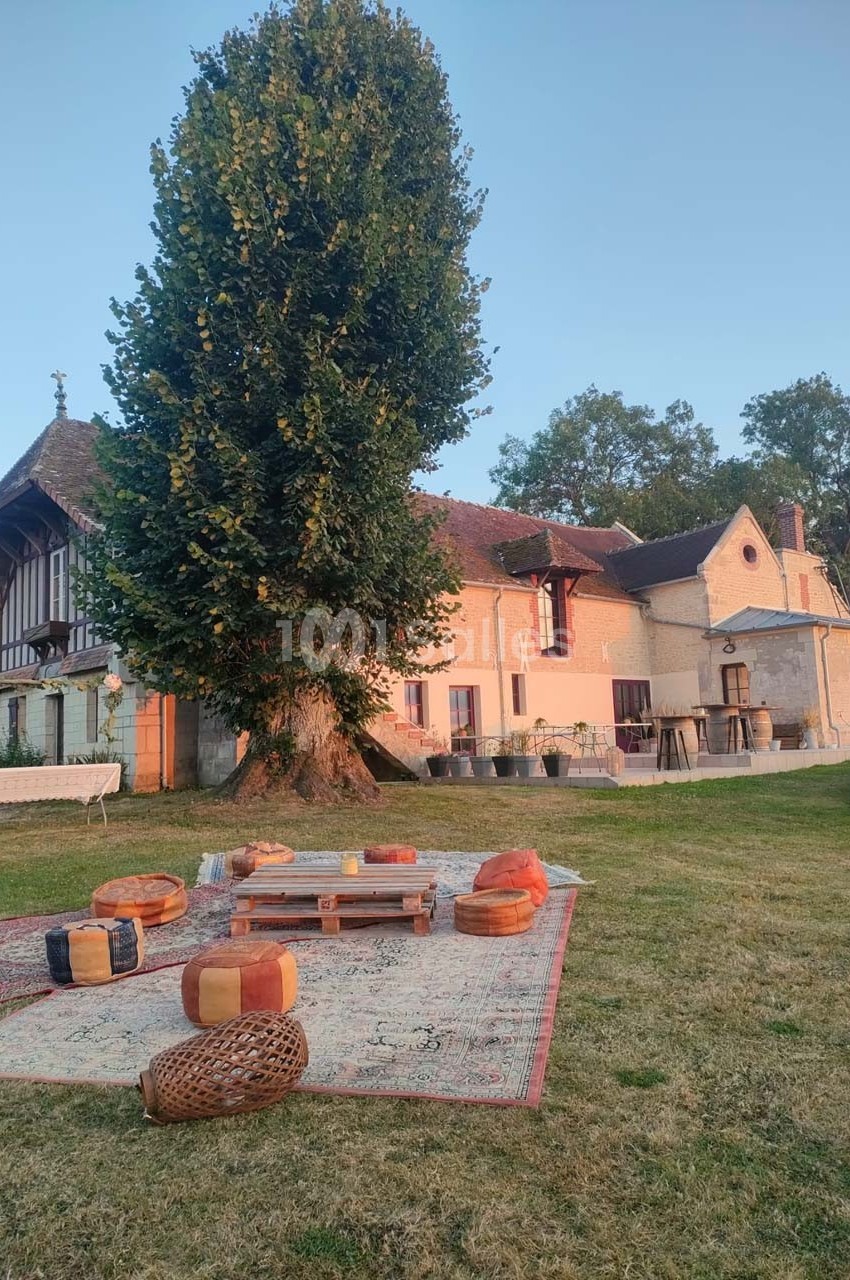 Espace extérieur avec tapis, coussins et table basse en bois devant une grande maison ancienne entourée de verdure.