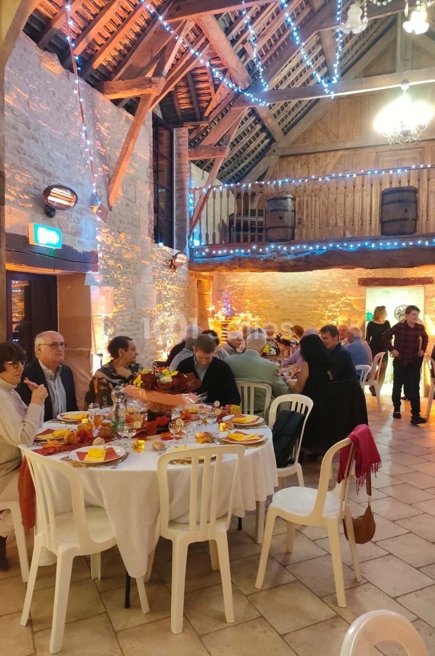 Salle rustique décorée avec des guirlandes lumineuses, des convives assis autour de tables dressées pour un repas convivial.