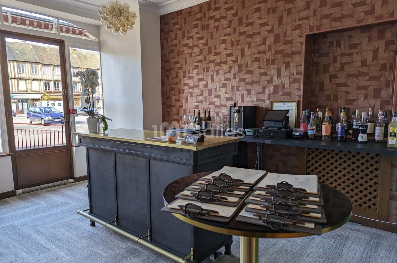 Intérieur d'un bar avec comptoir en bois sombre, étagères de bouteilles et table avec des menus sur pinces.