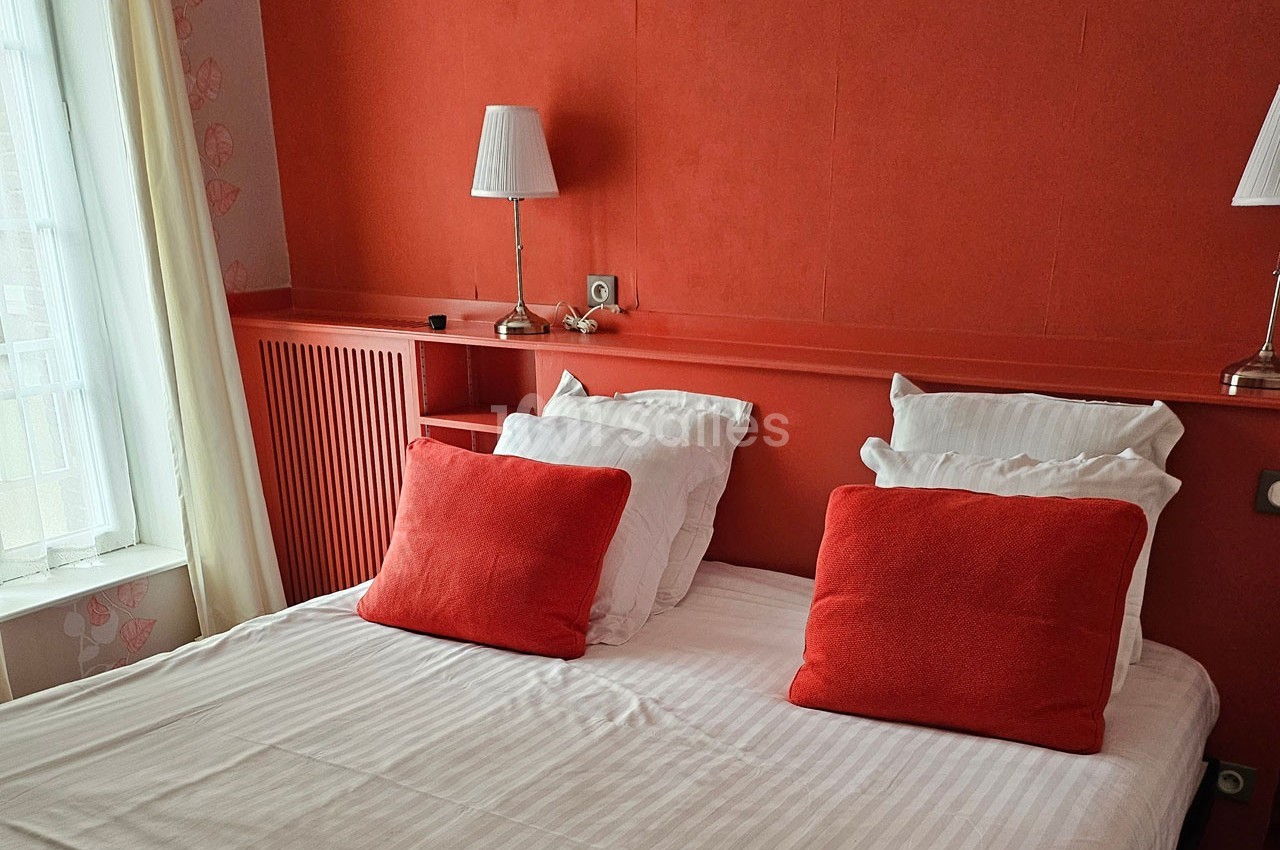 Lit double avec draps blancs, coussins rouges et lampes de chevet, dans une chambre aux murs rouges près d'une fenêtre.