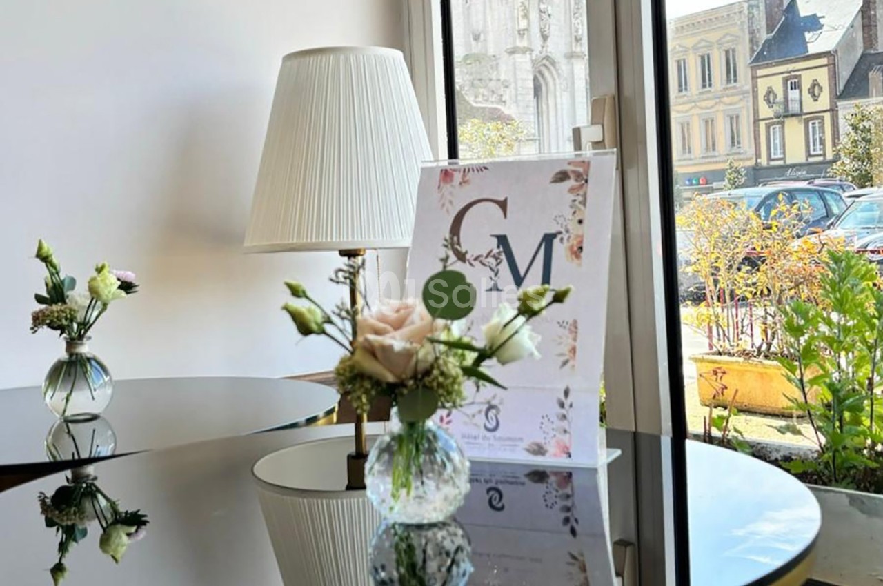 Vase avec des fleurs sur une table en verre, devant une lampe et une affiche, près d'une fenêtre donnant sur la rue.