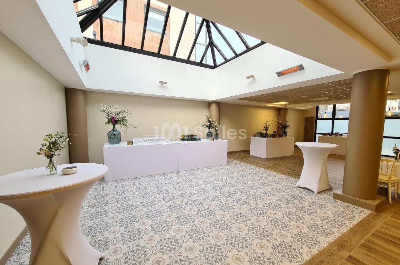 Salle lumineuse avec verrière, tables hautes habillées de nappes blanches, décorations florales et buffet en arrière-plan.