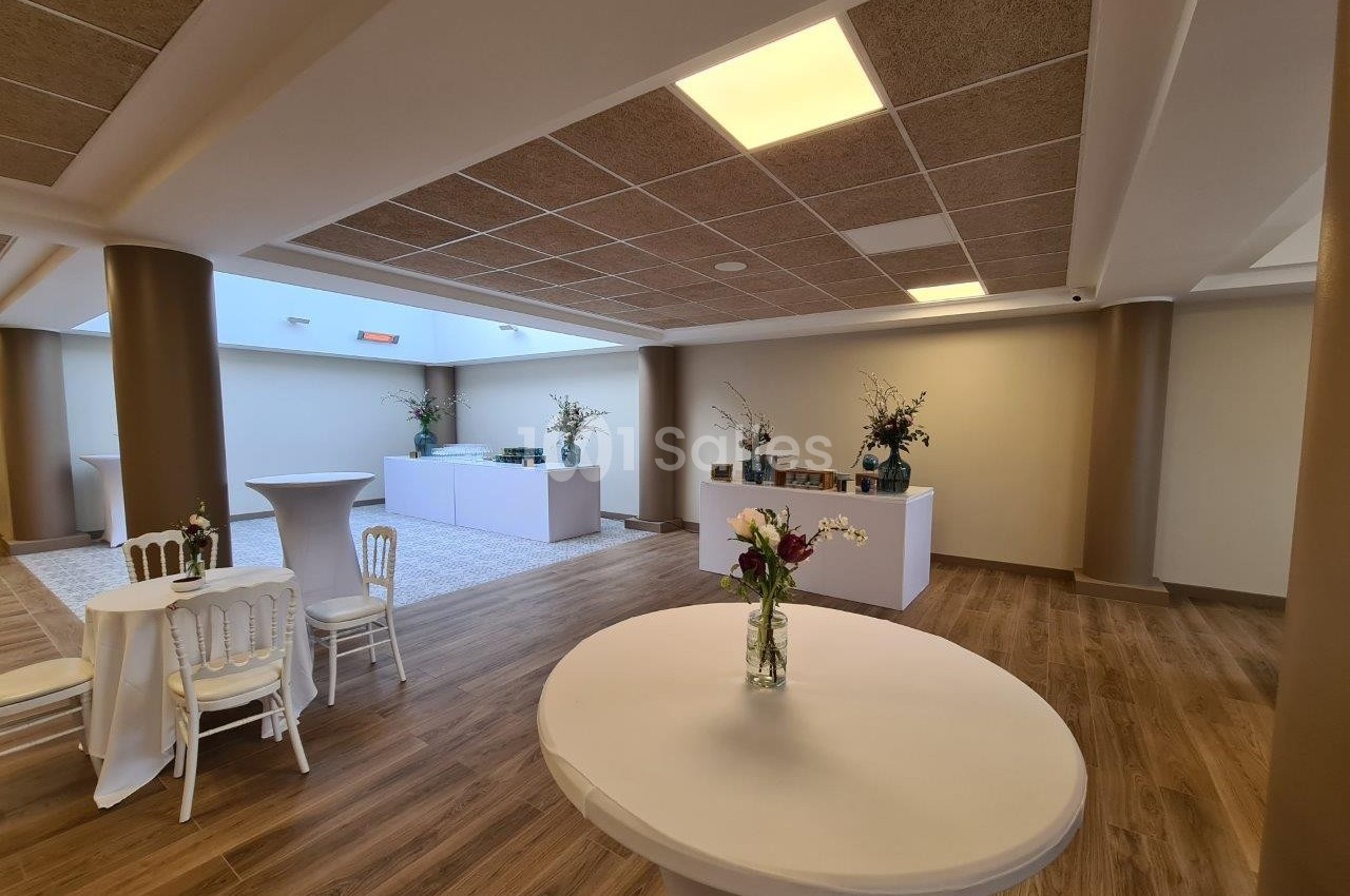 Salle lumineuse avec parquet, tables hautes nappées de blanc, chaises et décorations florales sur les tables.