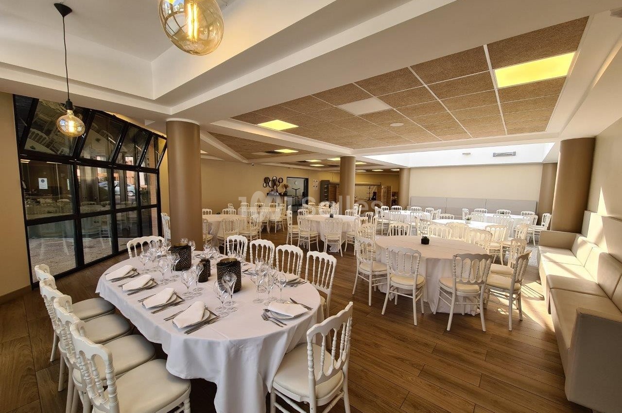 Salle de réception lumineuse avec tables rondes dressées, chaises blanches et décor sobre.