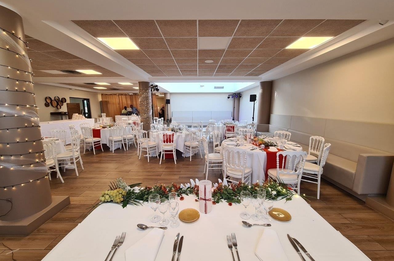 Salle de réception décorée pour un événement, avec tables dressées, chaises blanches et éclairage chaleureux.