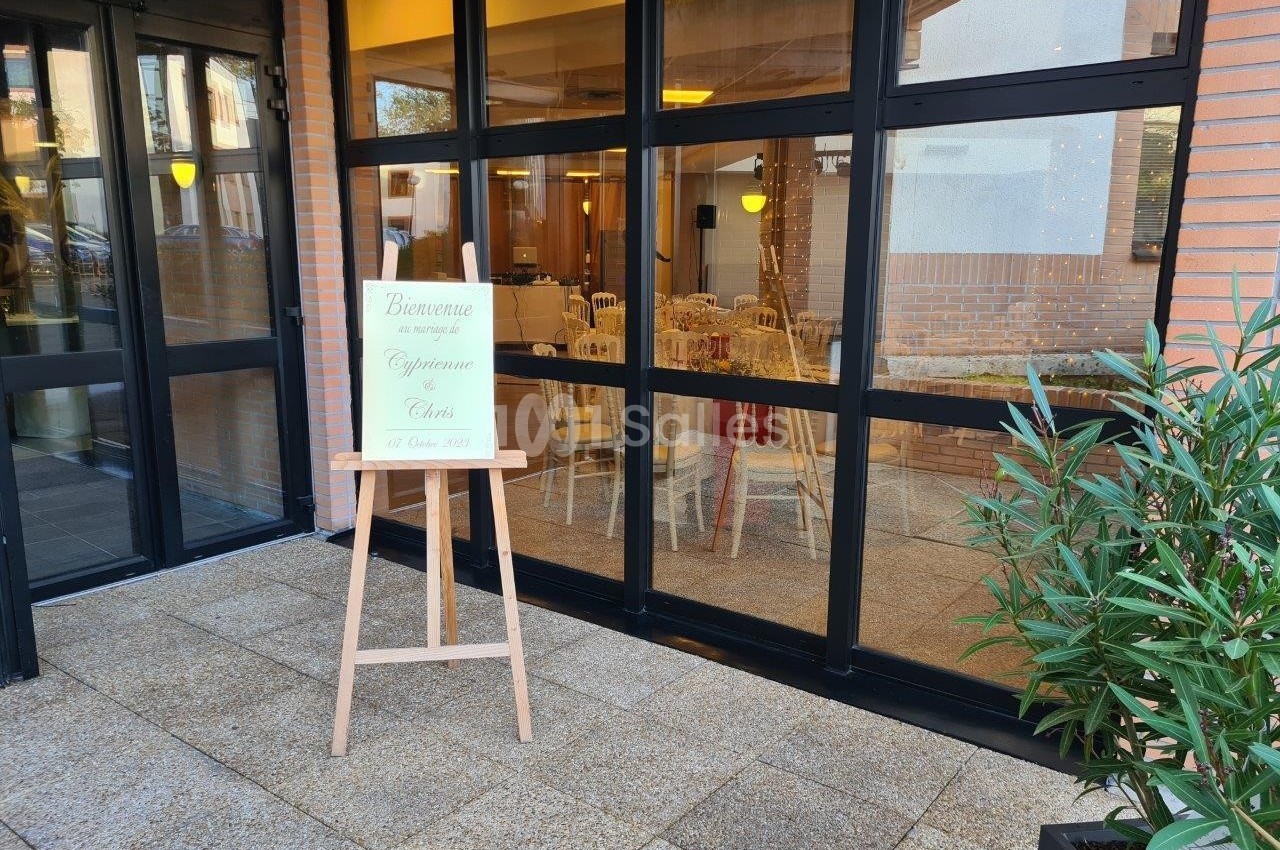 Panneau de bienvenue devant une salle vitrée aménagée pour un événement, avec des chaises et des tables décorées.