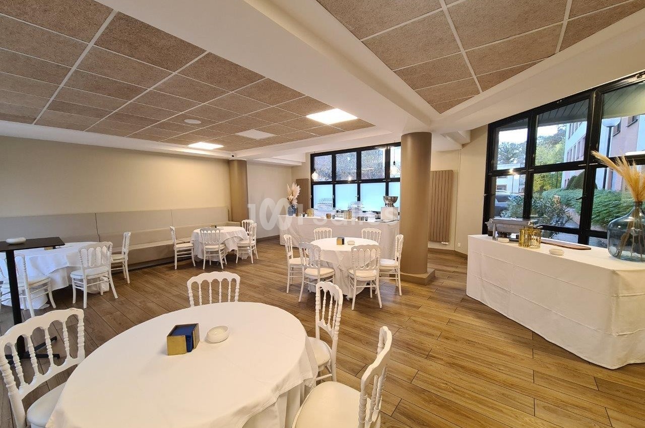 Salle lumineuse avec tables rondes dressées, chaises blanches et grandes fenêtres donnant sur l'extérieur.