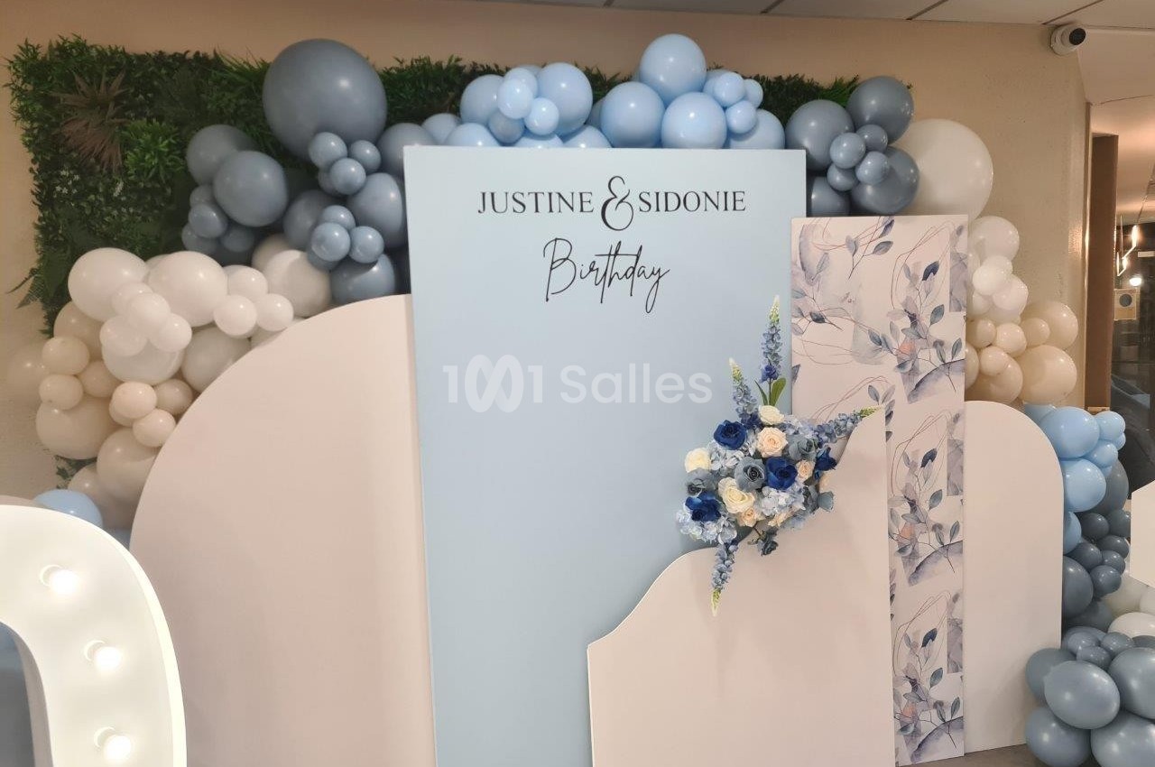 Mur décoratif avec ballons bleus et blancs, panneau ’Justine & Sidonie Birthday’ et arrangement floral bleu et blanc.