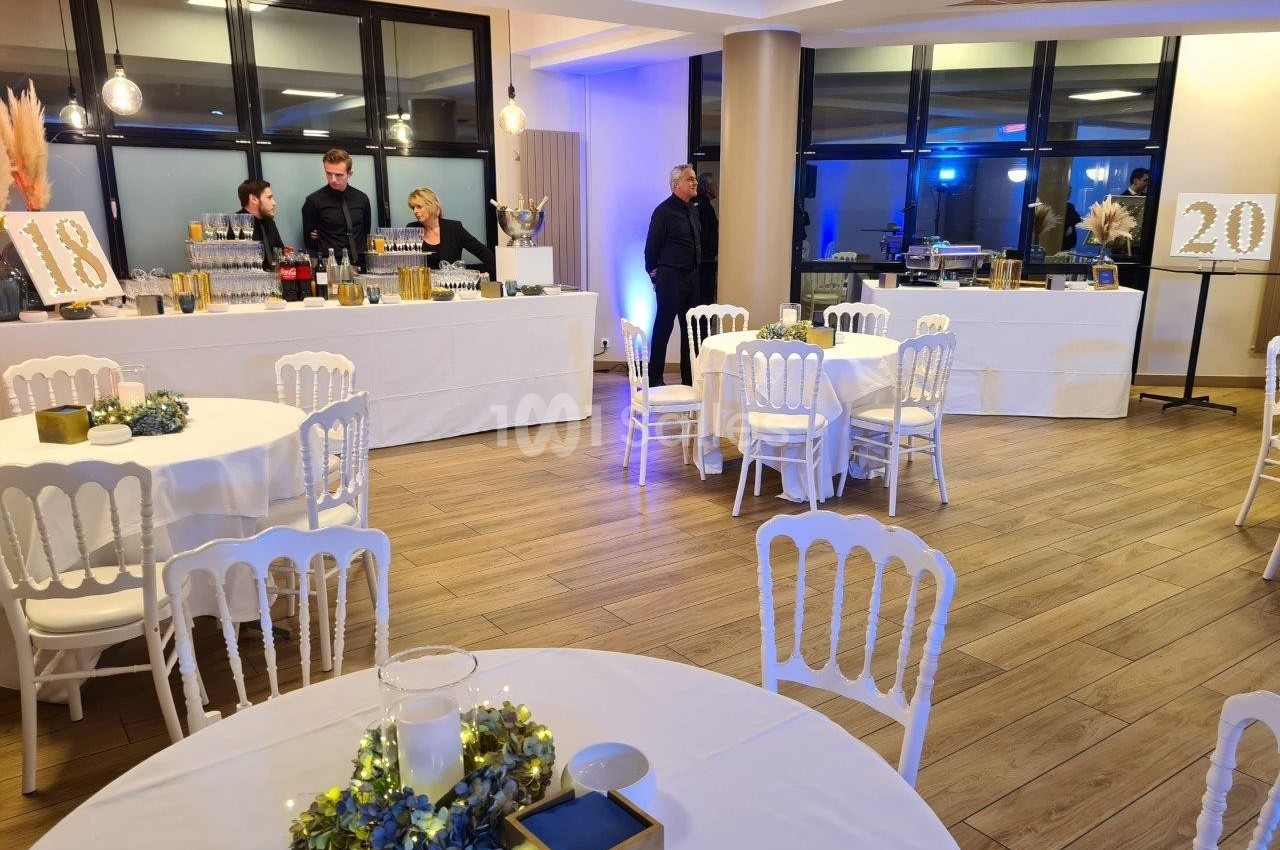 Salle décorée pour un événement avec tables rondes, chaises blanches et buffet en arrière-plan.