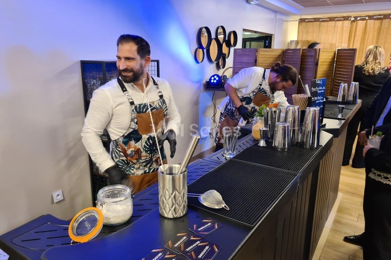 Deux barmans préparent des cocktails derrière un comptoir équipé d'ustensiles et de shakers dans un espace intérieur.