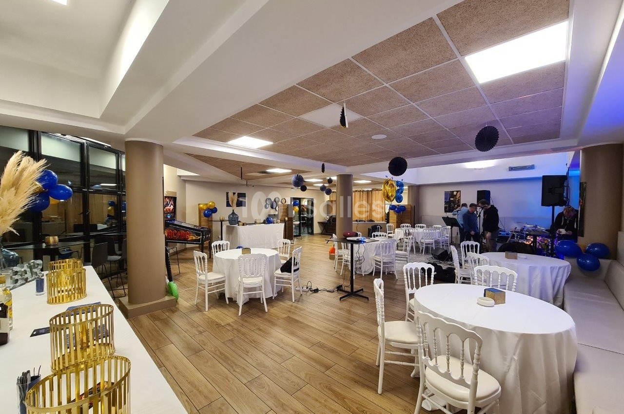 Salle décorée pour une fête avec tables rondes, chaises blanches, ballons bleus et noirs, et éclairage chaleureux.