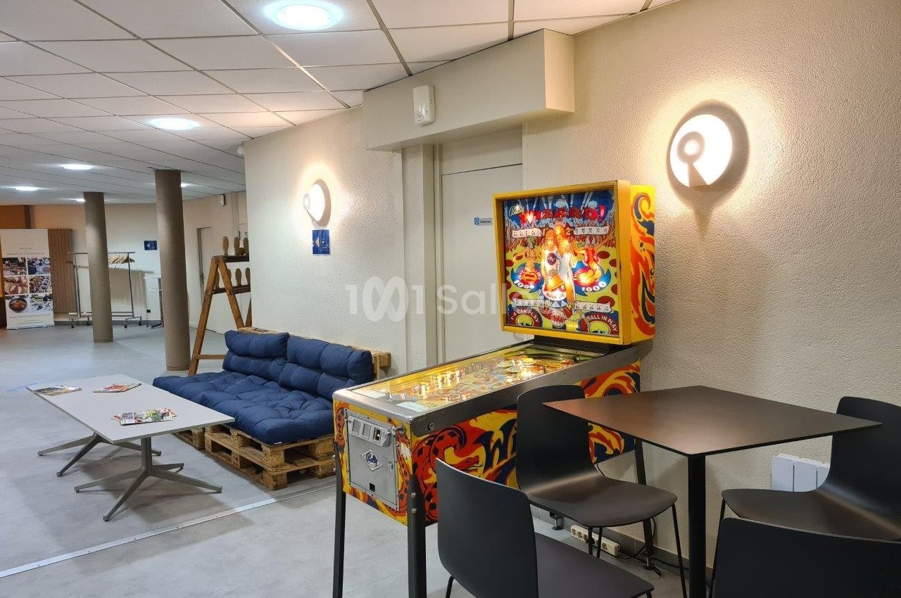 Flipper coloré dans un espace détente avec canapé en palettes, table basse et chaises hautes, éclairage mural.