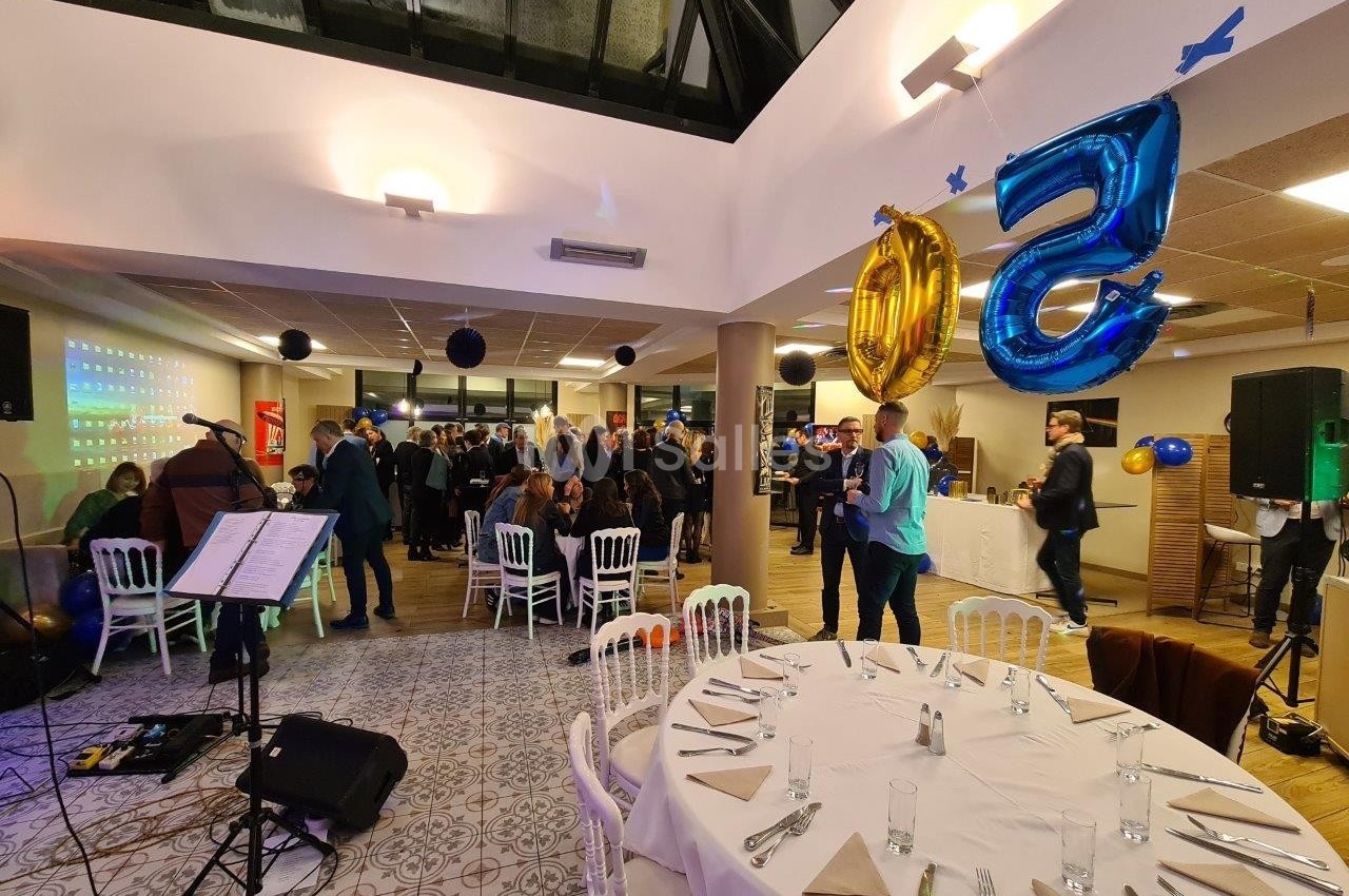 Salle décorée pour une fête avec des ballons ’50’, des invités debout et assis, et une scène musicale à gauche.