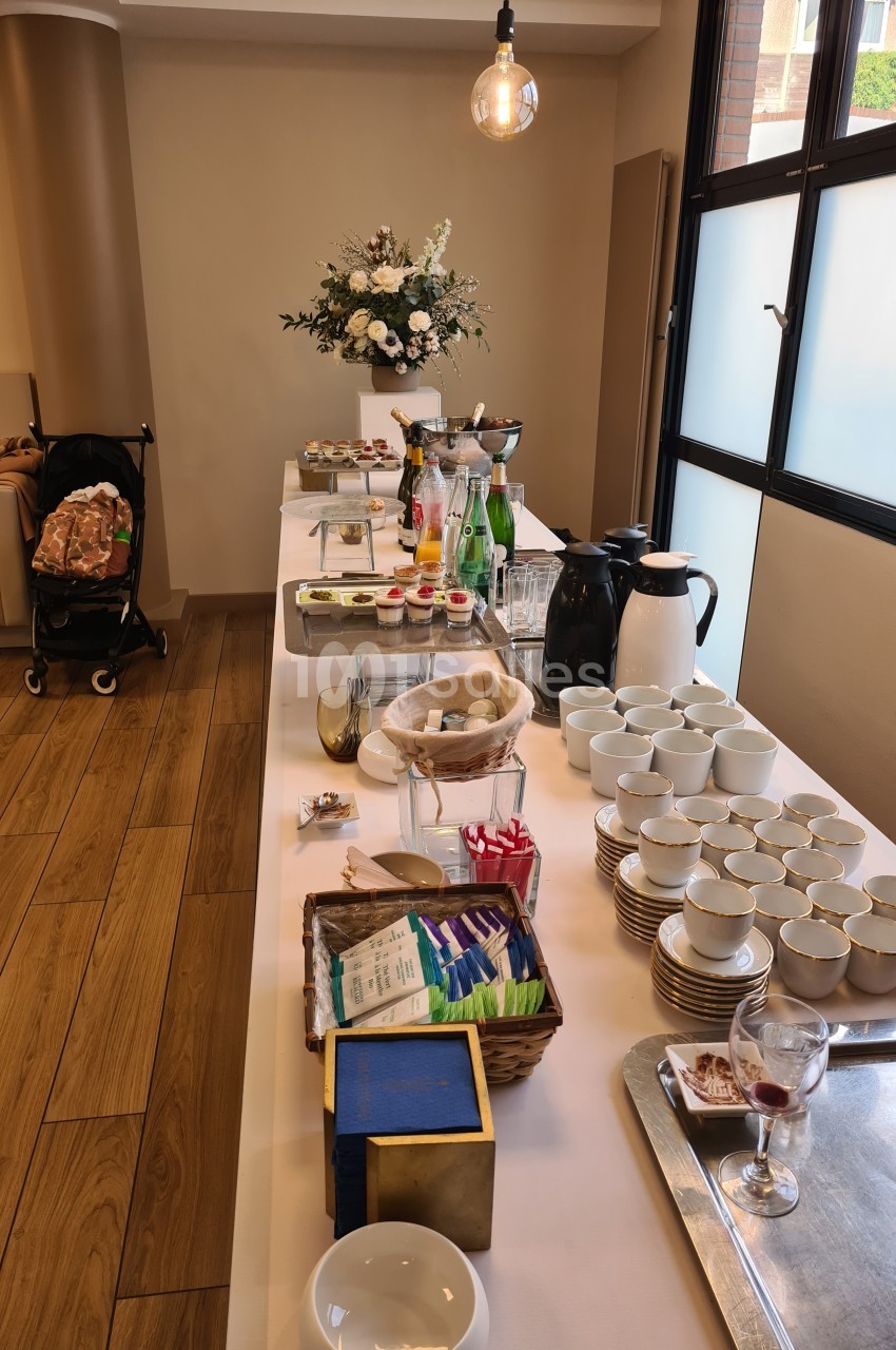 Table de buffet avec boissons, tasses, viennoiseries et fleurs, dans une salle lumineuse avec parquet et fenêtres.