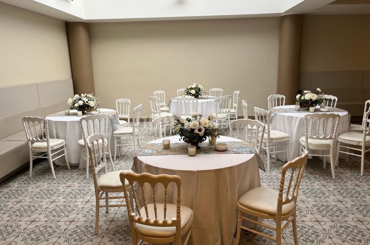 Salle lumineuse avec tables rondes décorées de nappes blanches et bouquets de fleurs, entourées de chaises élégantes.