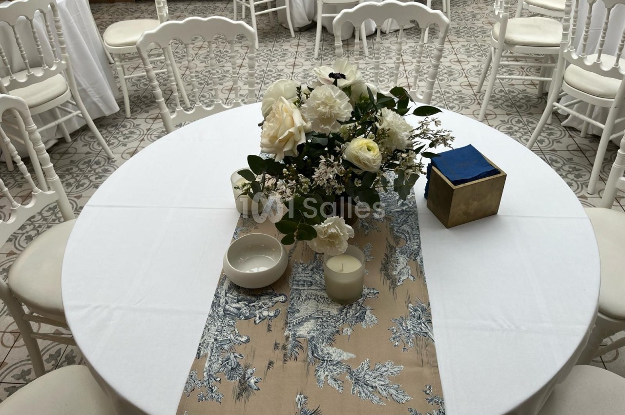 Table ronde décorée avec une nappe blanche, un chemin de table beige à motifs, des fleurs blanches et des bougies.