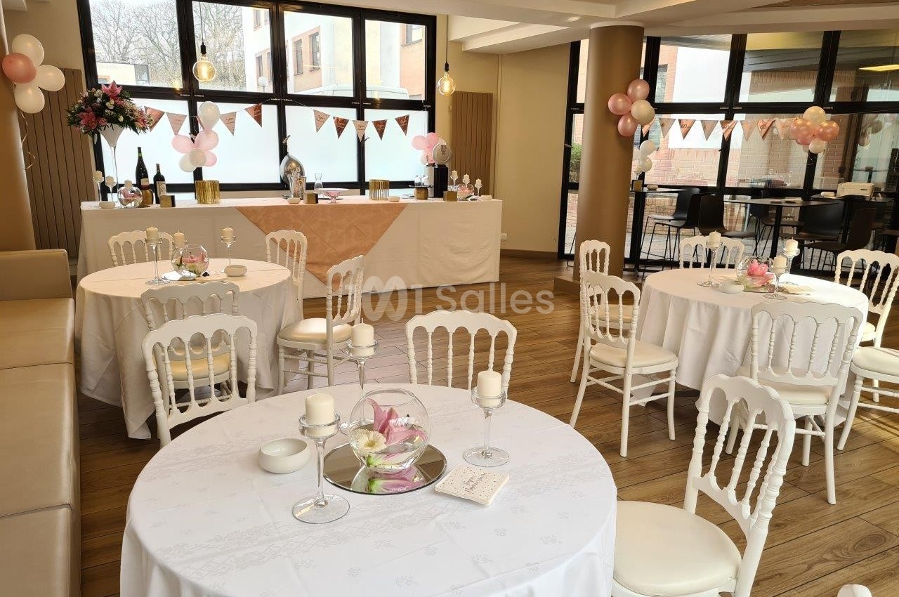 Salle décorée pour une réception avec tables rondes dressées, guirlandes et ballons roses en arrière-plan.