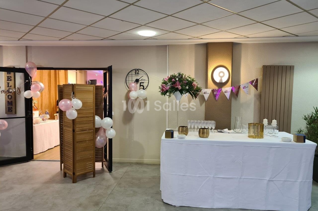 Salle décorée pour un événement avec table dressée, ballons, guirlandes et fleurs près d'une cloison en bois.