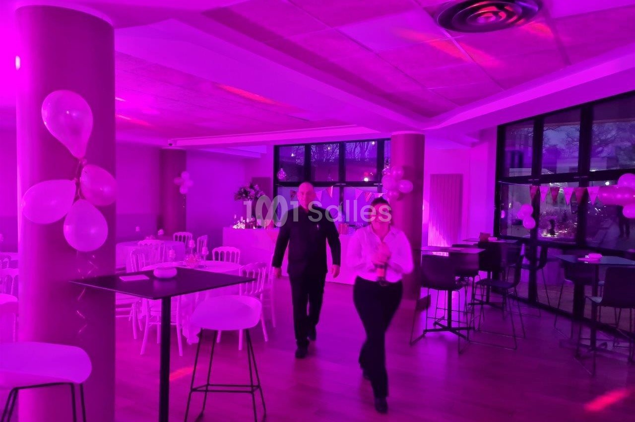 Salle décorée avec des ballons roses, éclairage violet, deux personnes marchant au centre, tables hautes et chaises…