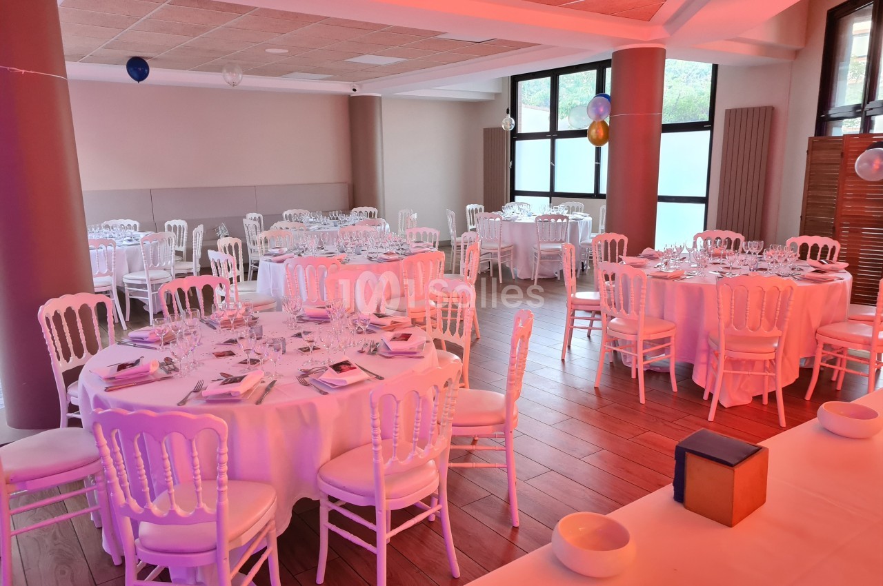 Salle de réception décorée avec des tables rondes dressées, chaises blanches et éclairage rosé.