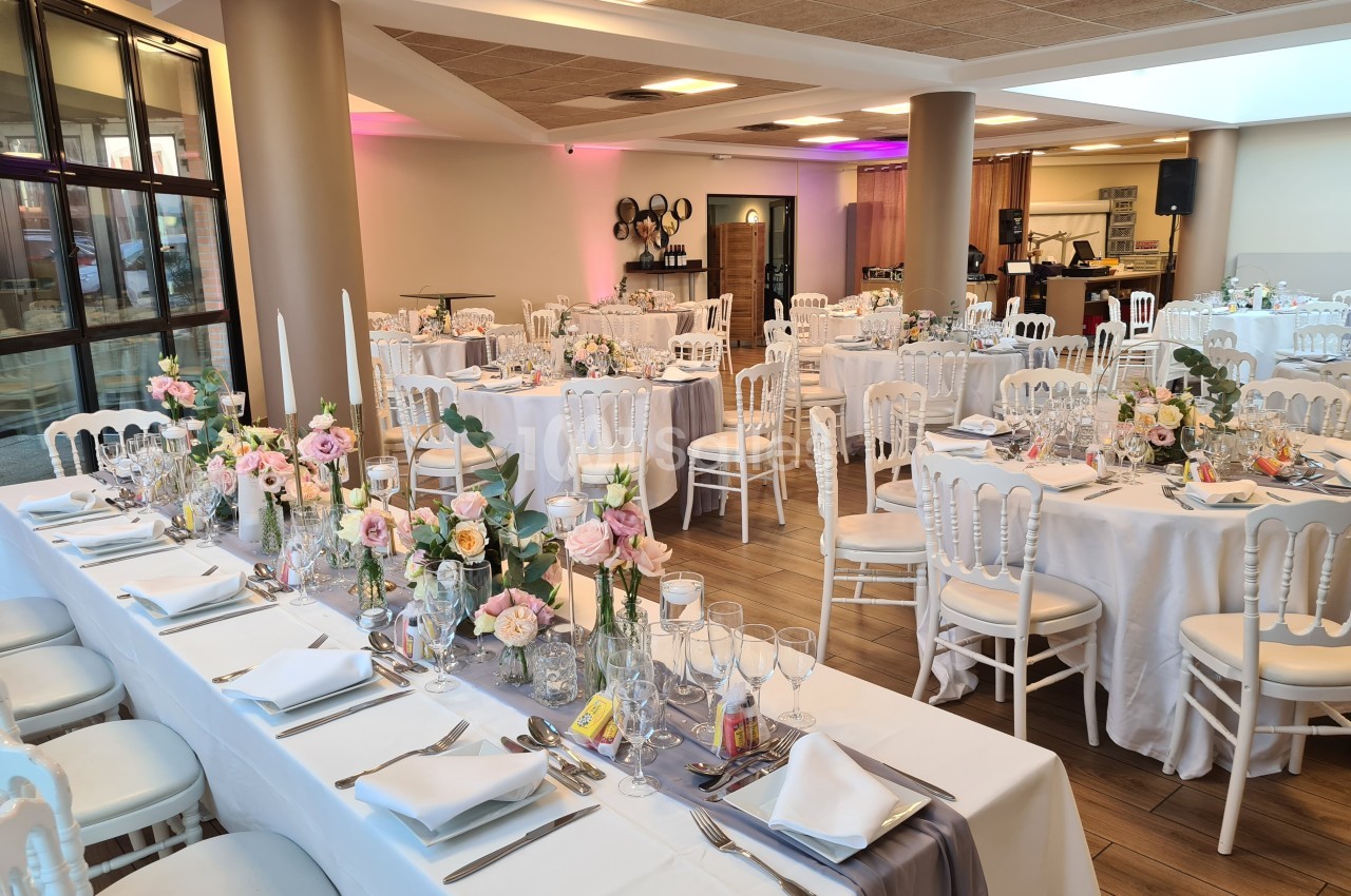 Salle de réception décorée avec des tables dressées, nappes blanches, fleurs et chaises blanches dans un cadre lumineux.