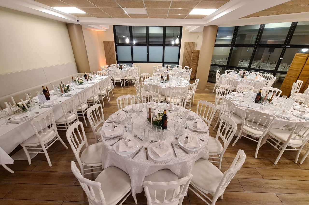 Salle de réception avec tables rondes et rectangulaires dressées, nappes blanches et chaises en bois clair.