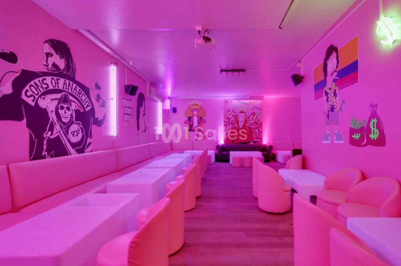 Salle aux murs décorés de fresques colorées, éclairée par des néons roses, avec des tables et fauteuils blancs.