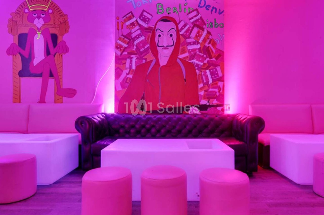 Salon avec canapé en cuir noir, tables blanches, poufs roses et mur décoré de peintures colorées sous éclairage rose.