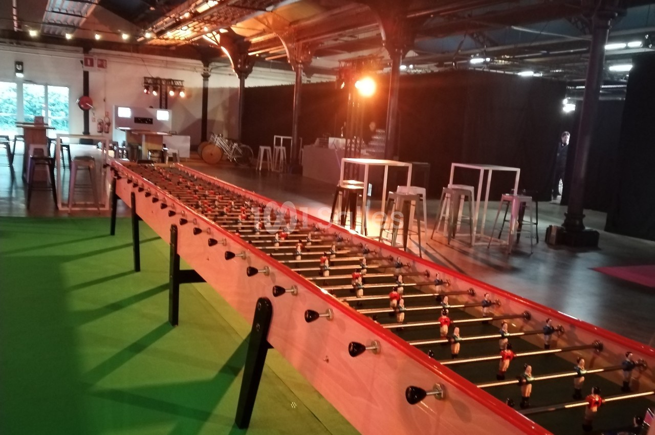 Baby-foot géant installé dans un espace intérieur avec éclairage tamisé et tables hautes en arrière-plan.