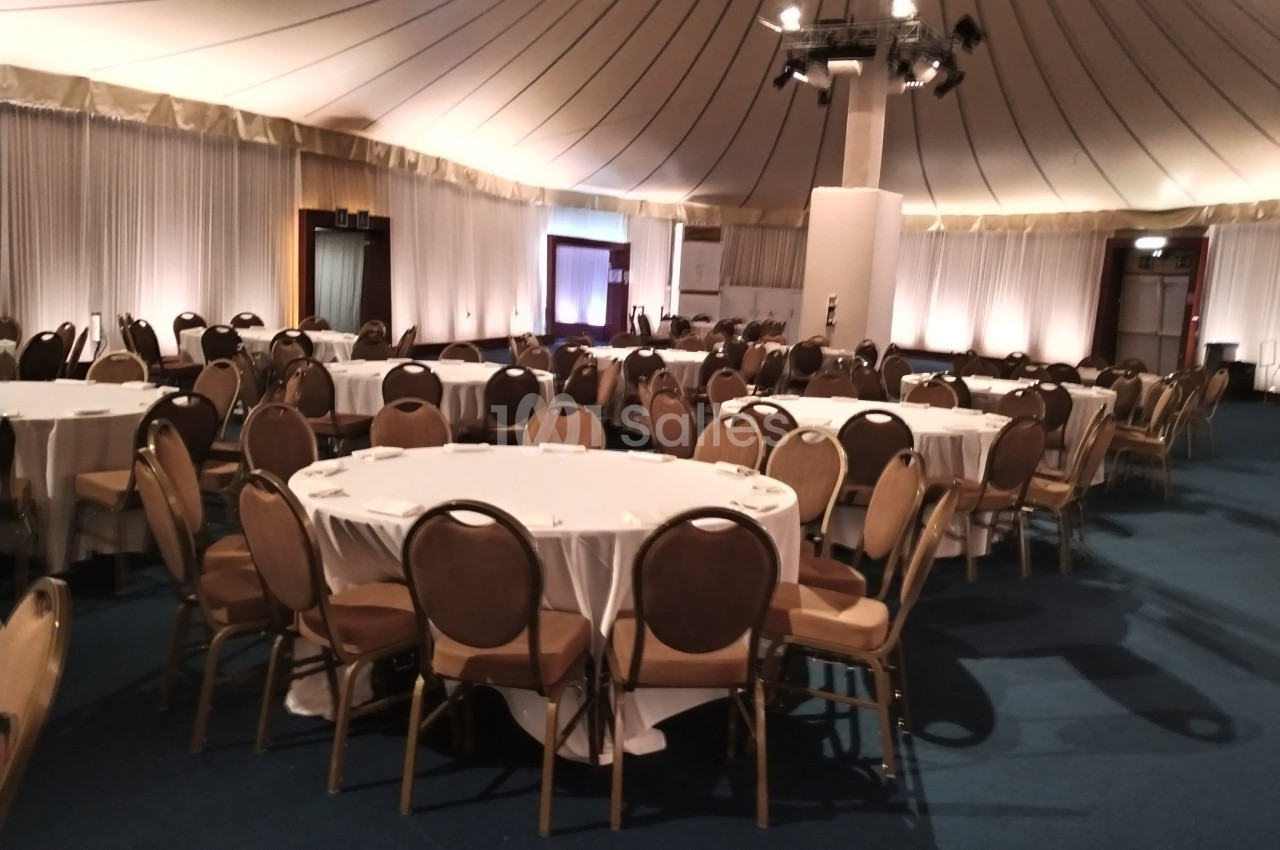 Salle de réception avec des tables rondes dressées de nappes blanches et entourées de chaises marron.