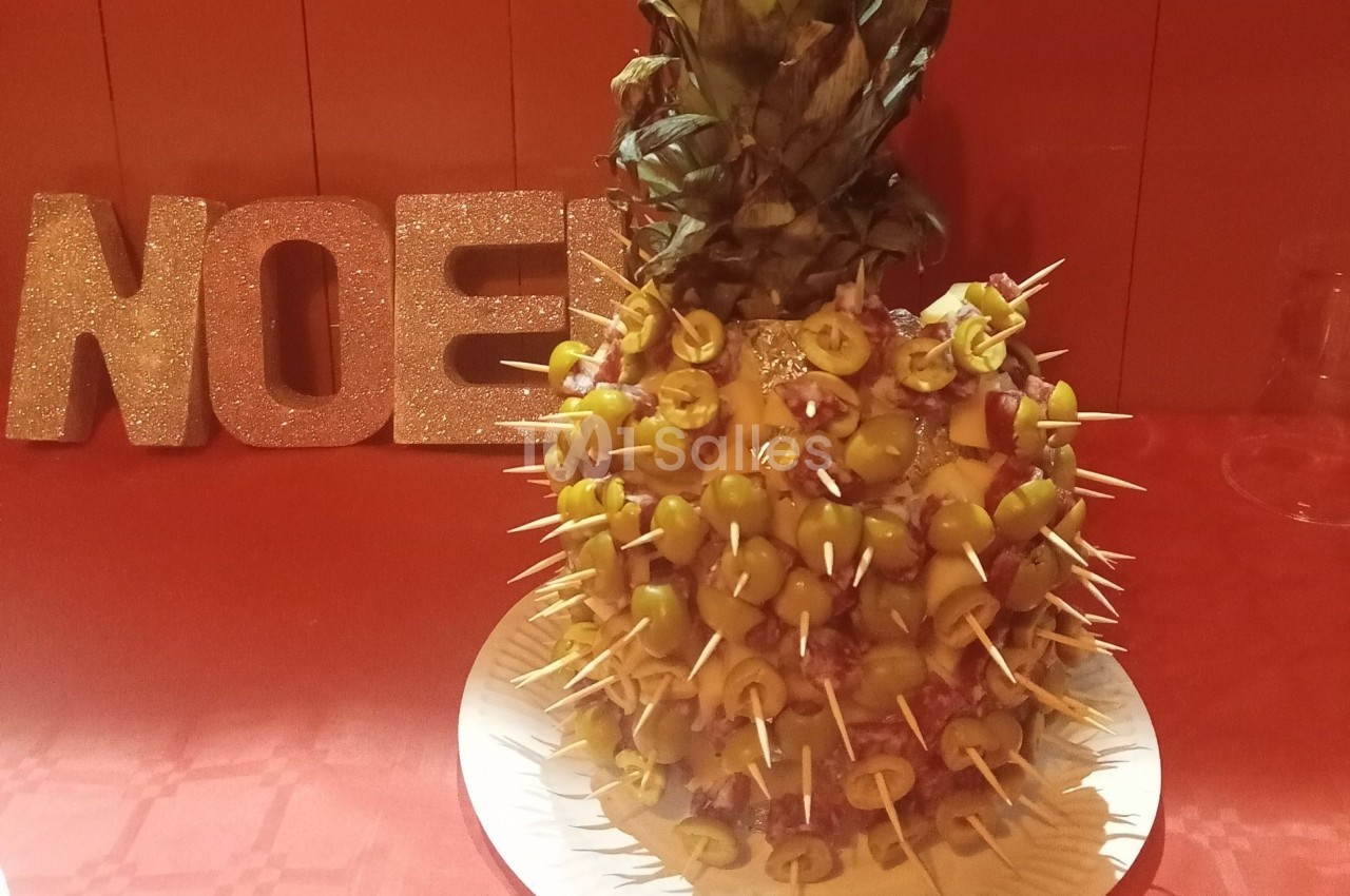 Ananas décoré de brochettes d'olives et de charcuterie, posé sur une table rouge avec des lettres ’NOEL’ scintillantes en…