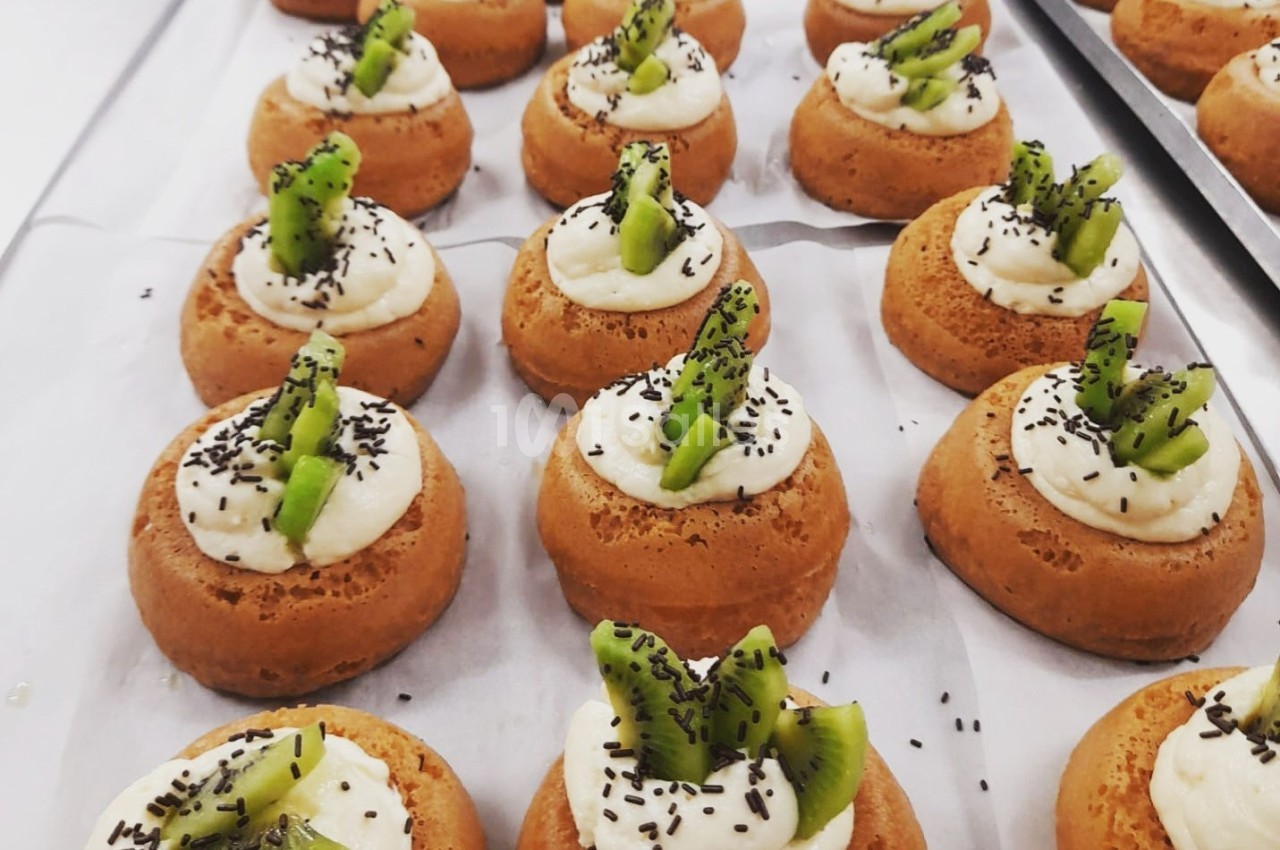 Petits gâteaux individuels garnis de crème et de morceaux de kiwi, décorés de graines noires sur du papier cuisson.