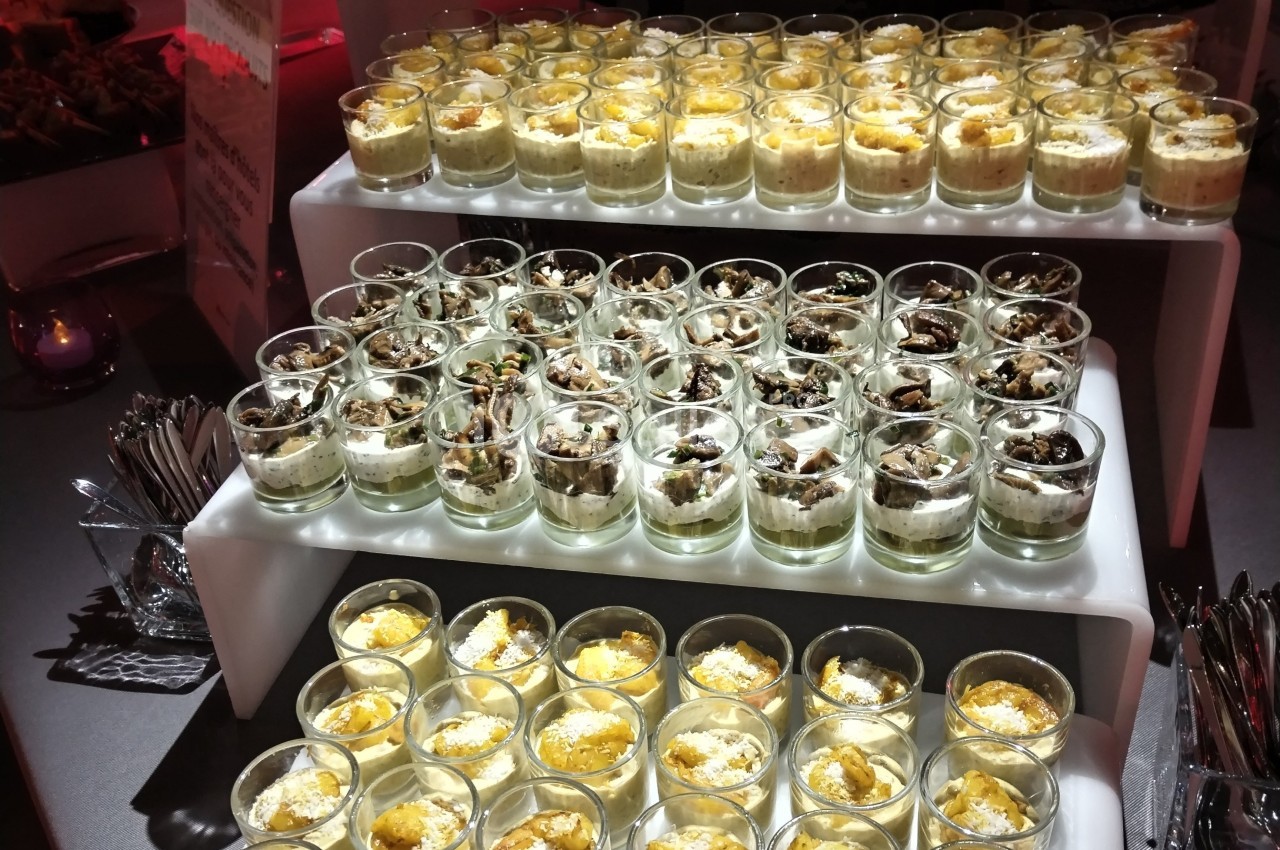 Buffet de verrines variées disposées sur des étagères blanches, éclairées par une lumière tamisée.