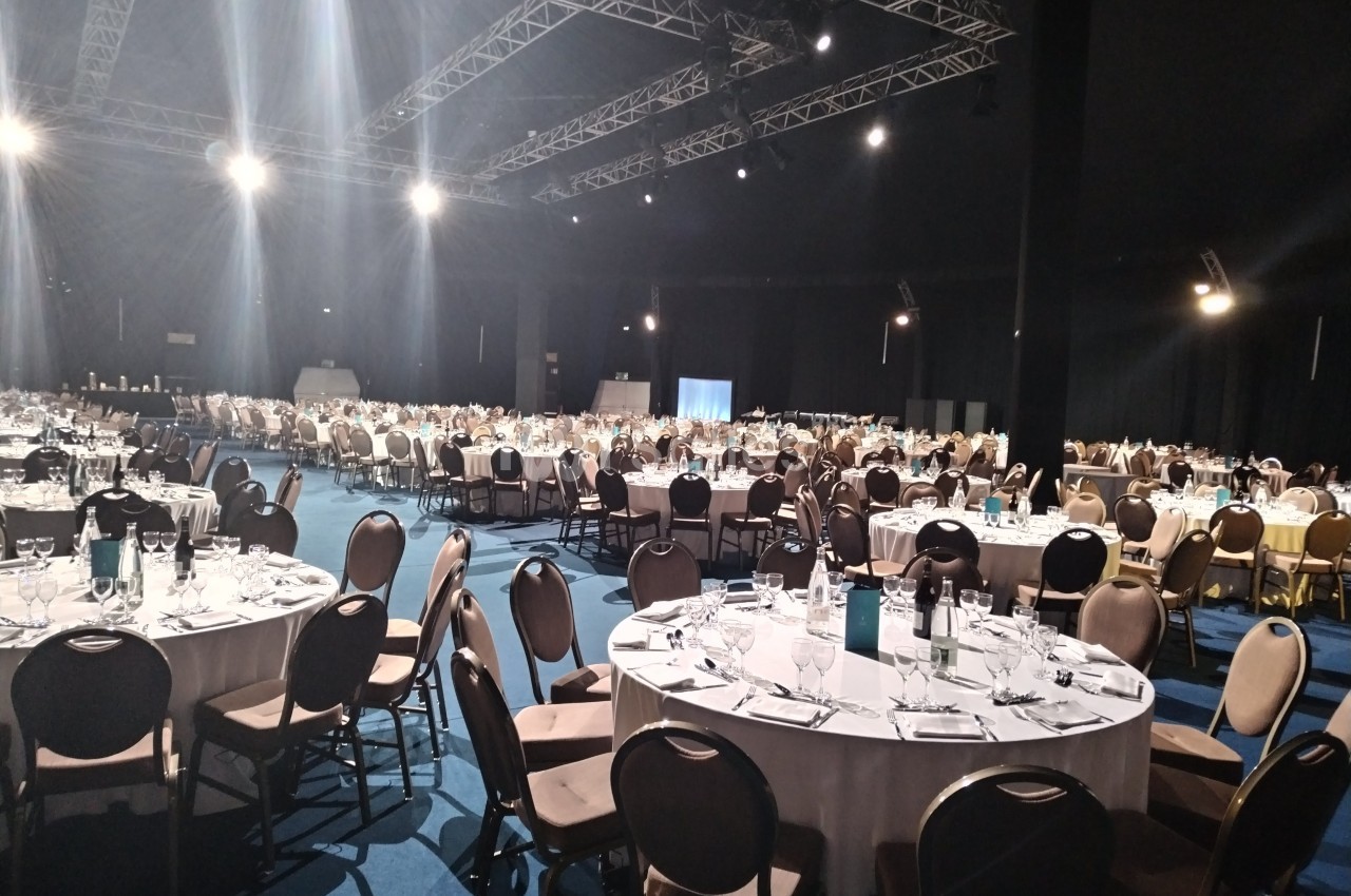Salle de réception avec de nombreuses tables rondes dressées, éclairée par des projecteurs au plafond.