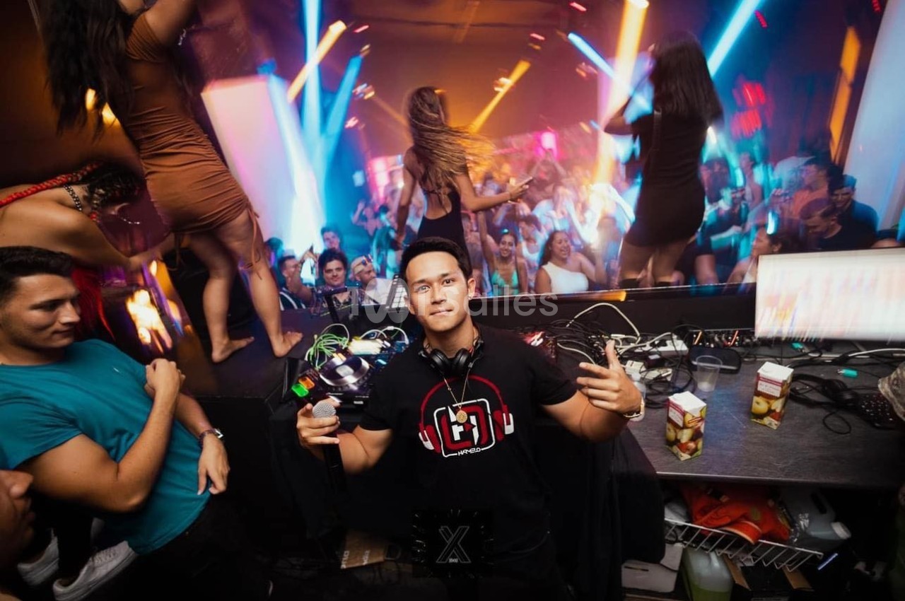 Un DJ pose devant son équipement dans une boîte de nuit animée avec des lumières colorées et une foule dansante.