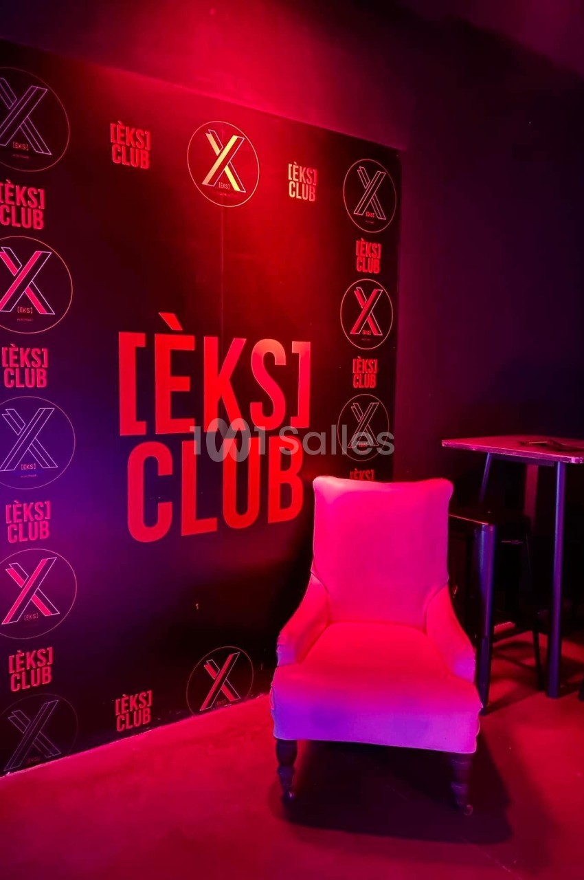 Fauteuil rose devant un mur noir avec le logo ’[ÈKS] CLUB’ illuminé par une lumière rouge.