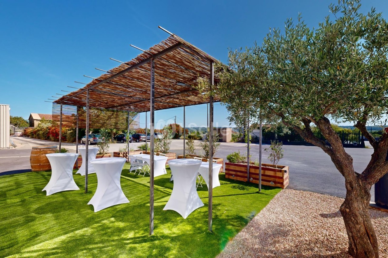 Espace extérieur avec tables hautes sous une pergola en bois, entouré de végétation et d'un sol en gazon artificiel.