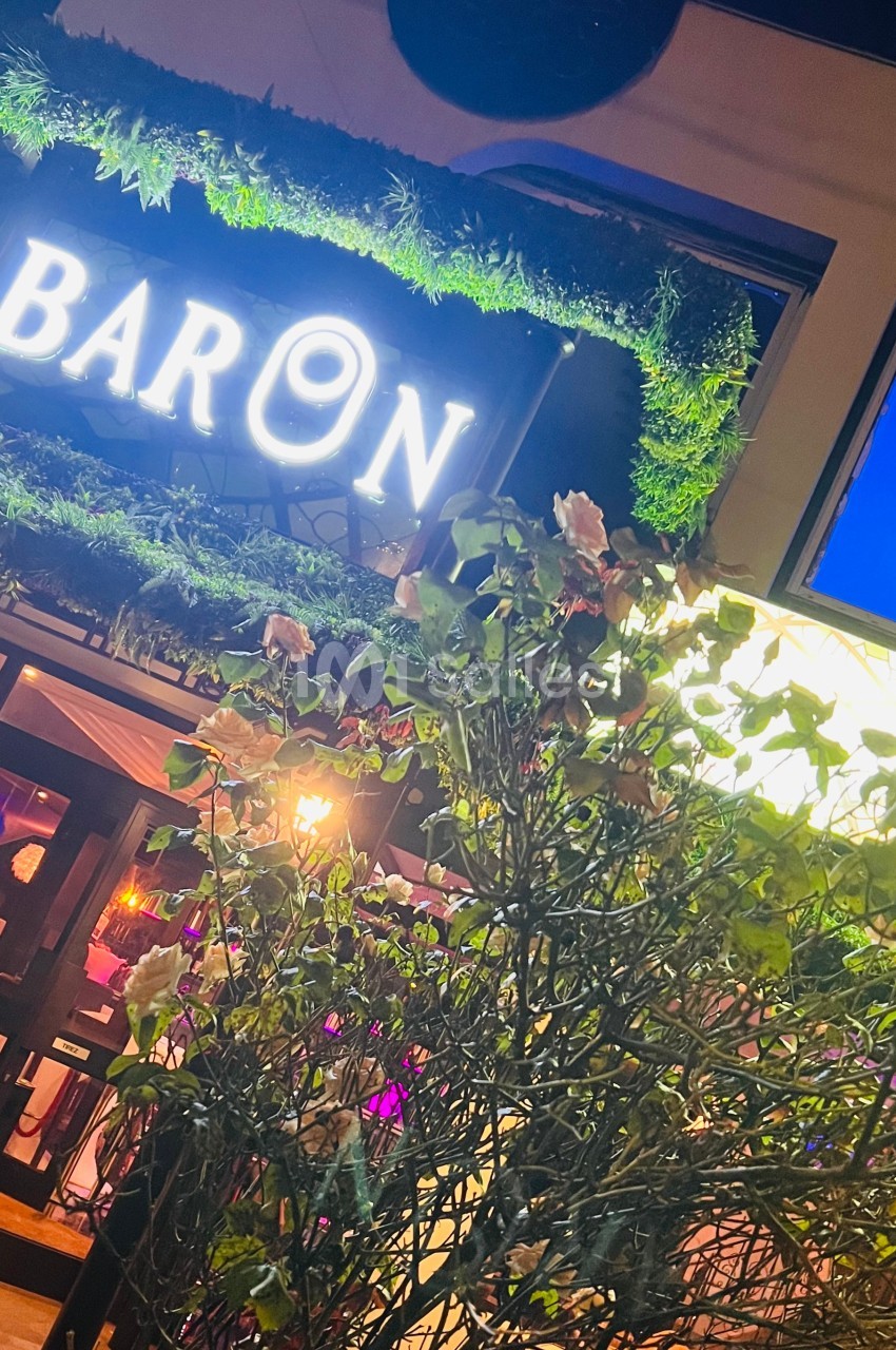 Enseigne lumineuse ’Baron’ entourée de verdure, avec des roses au premier plan et une façade éclairée en arrière-plan.