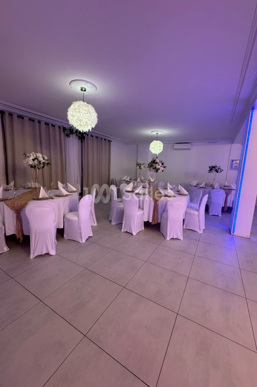 Salle de réception décorée avec des tables nappées, chaises blanches et centres de table floraux sous des luminaires…