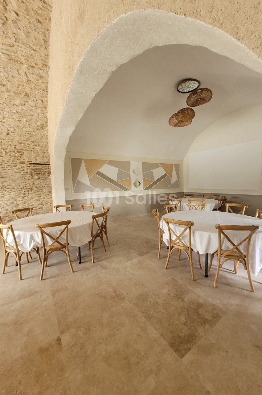 Salle lumineuse avec murs en pierre, tables rondes couvertes de nappes blanches et chaises en bois clair.
