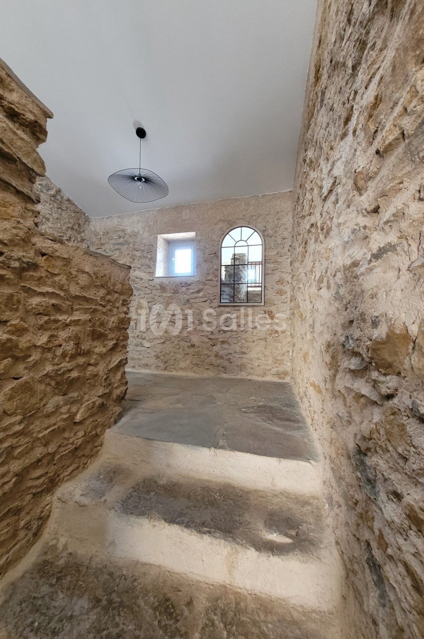 Escalier en pierre dans un intérieur aux murs en pierre apparente, éclairé par une fenêtre et une suspension moderne.