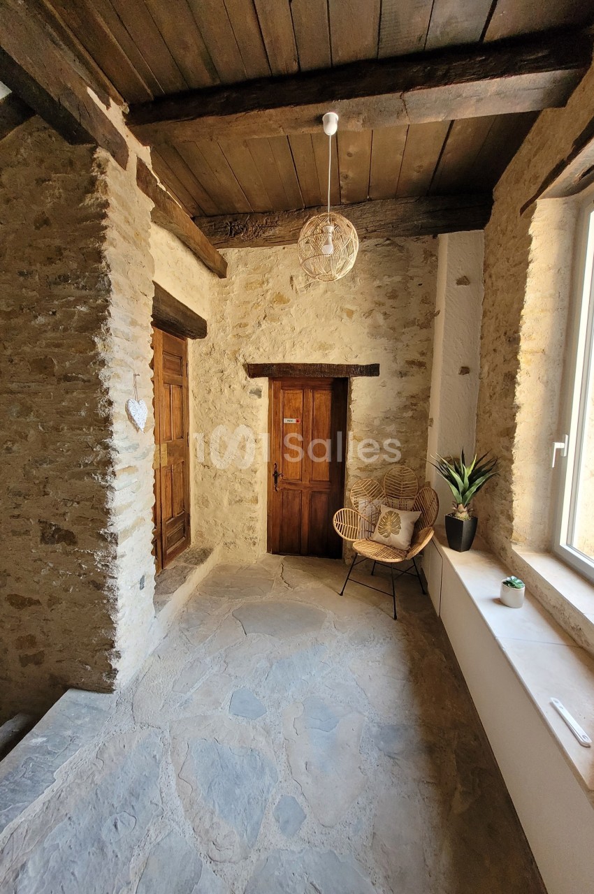 Couloir en pierre avec plafond en bois, porte en bois, fauteuil en rotin, plante et lumière naturelle provenant d'une…