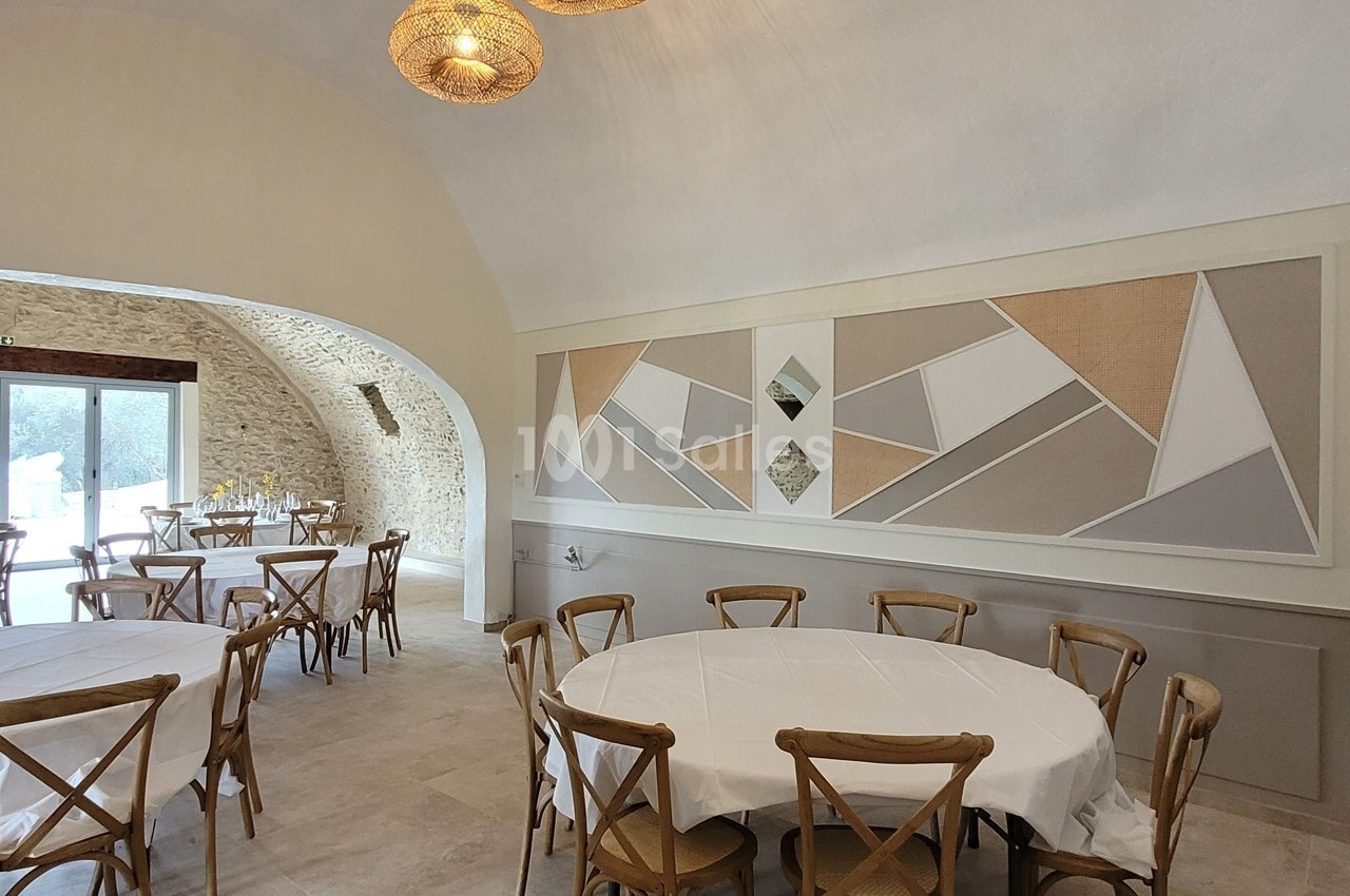 Salle lumineuse avec tables rondes dressées, chaises en bois, murs en pierre et décoration murale géométrique.