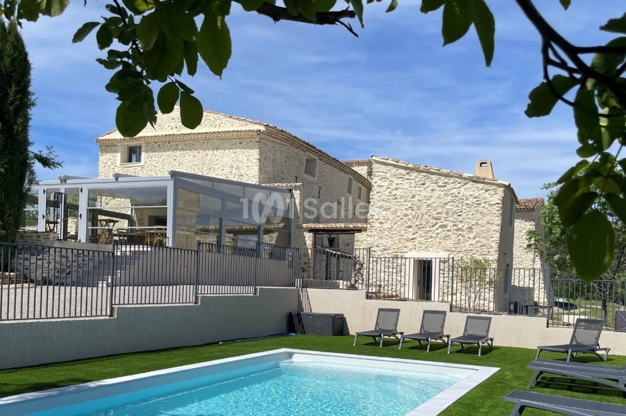 Maison en pierre avec terrasse vitrée, piscine et transats, entourée de végétation sous un ciel bleu.